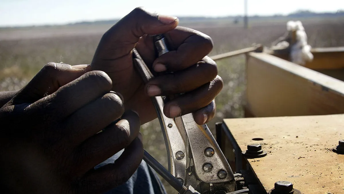 A close-up shot of two hands on a cotton module maker with farmland visible in the background.