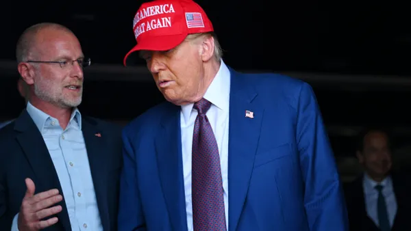 Brendan Carr stands to the left of Donald Trump, who is wearing a red hat that says "Make America Great Again."