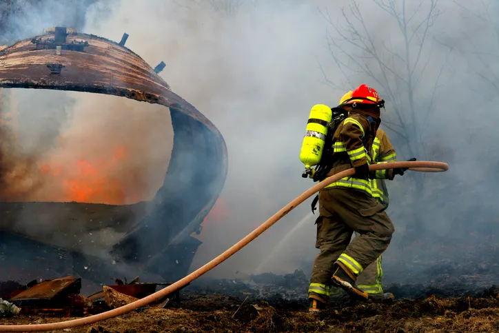 Two people in firefighting suits carry a hose outside. Fire and smoke is billowing in front of them.