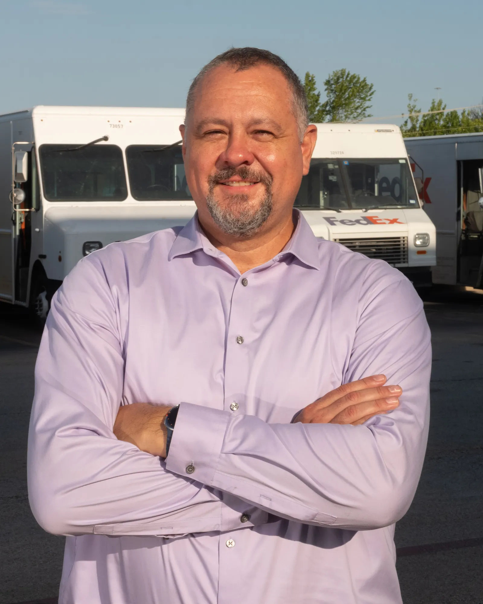 A person with a light purple shirt stands with crossed arms in front of a row of parked white FedEx trucks.