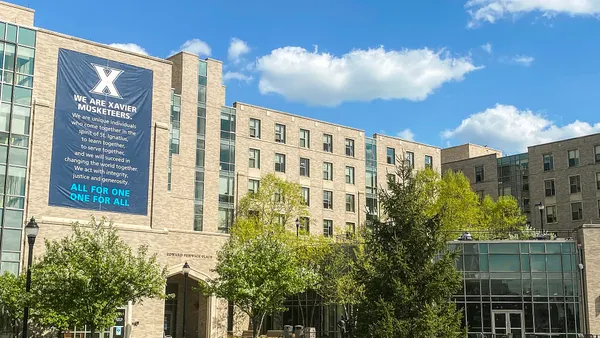 White brick building with large blue sign with header saying "We are Xavier Muskateers."