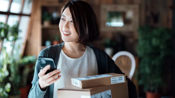 A women in a cardigan sweater and white shirt smiles while holding a phone in her right hand and two packages in her left.