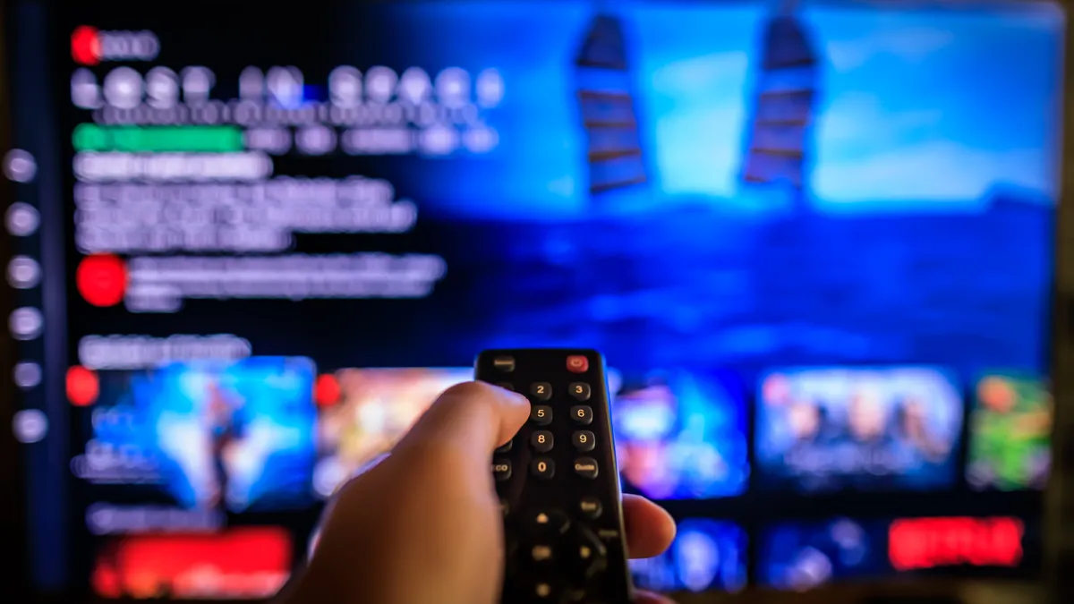 a person holds a remote in front of a TV showing a blurred out streaming service interface.