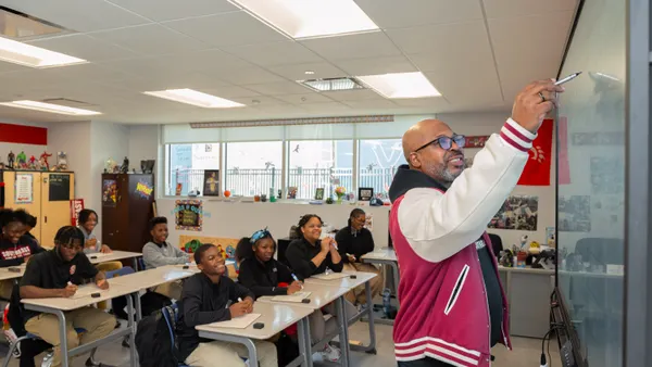 An adult is standing in the front of a classroom, writing on a whiteboard. The person's back is to a classroom of students sitting at individual desks and looking ahead.