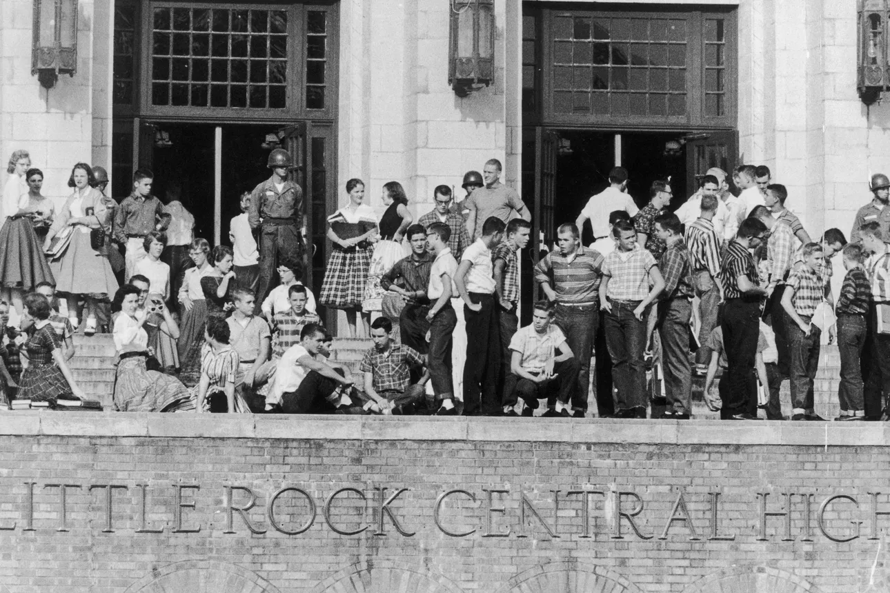 A line of White students stand outside of Little Rock Central High School, with the National Guard standing behind them, blocking the entrance to the school.