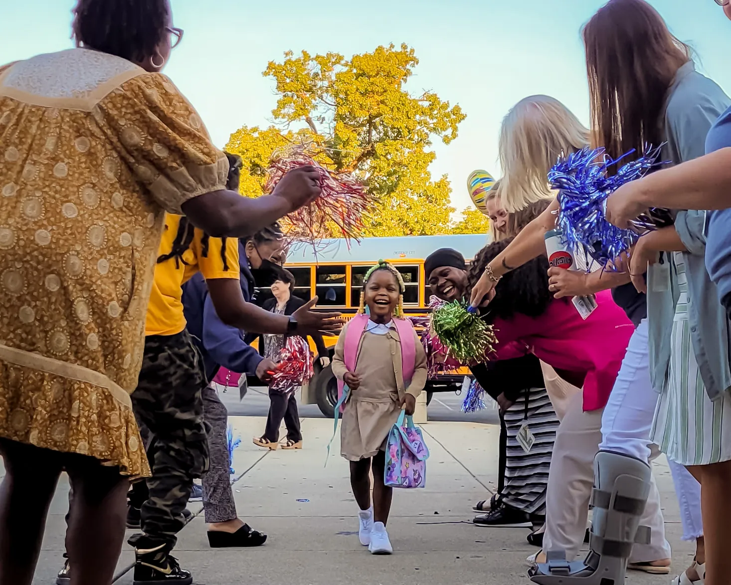 A young student walks between two lines of people holding pom-poms and high-fiving. A yellow school bus is in the background