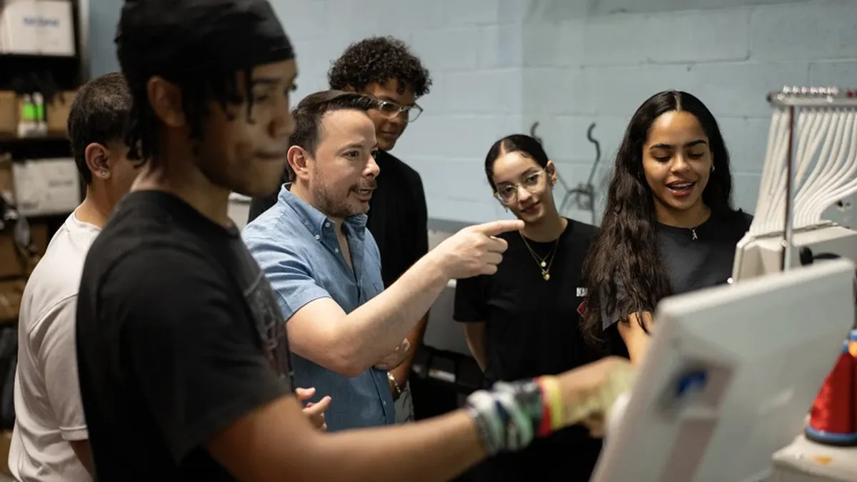 A principal is pictured standing among students, speaking to them as they use an embroidery machine in a commercial art course.