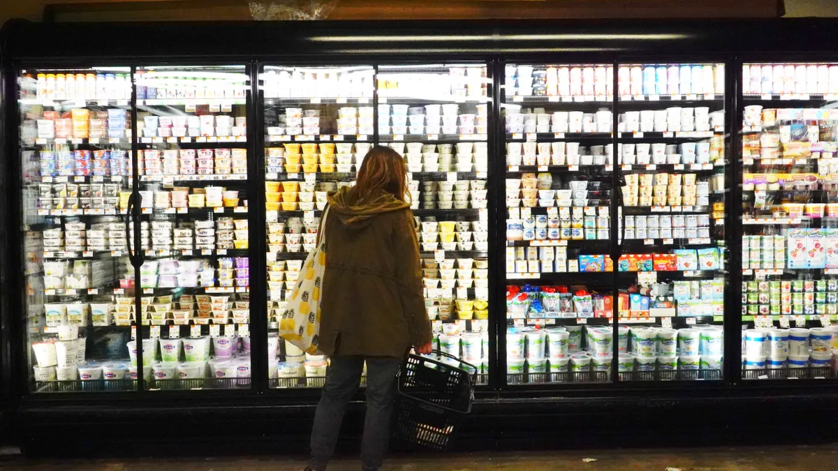 A person holding a shopping cart stands in front of a row of refrigerators stocked with food at a grocery store.