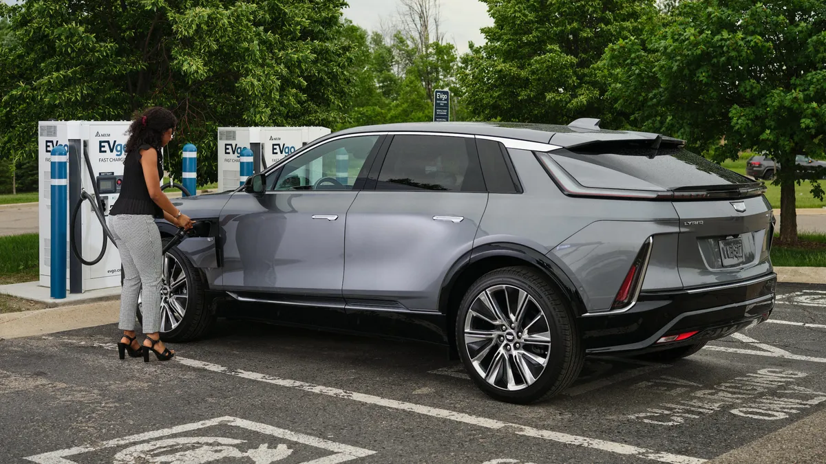 A gray four-door crossover vehicle parked in an electric vehicle charging stall with a woman holding a charging cable to the car.