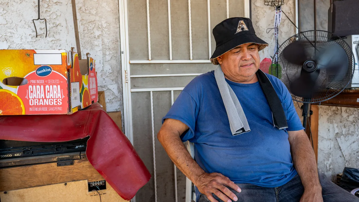 A person with a hat on and towel around neck sits on a porch in front of a fan.