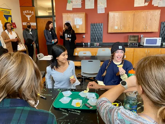 Students are seated at a table in a classroom. Two are holding lemon slices while two others have their backs to the camera.