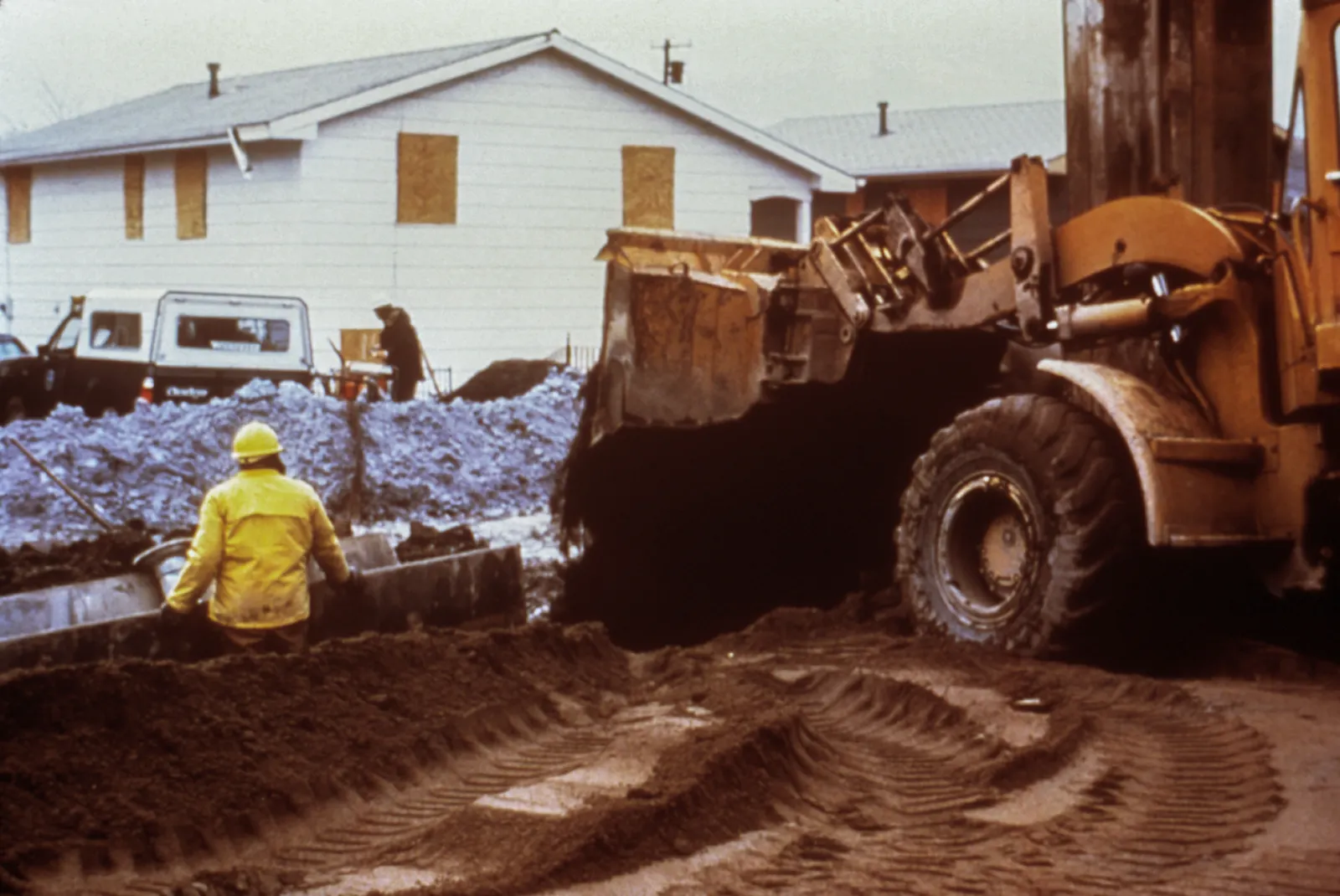 A person in safety gear stands in a trench, next to an excavator, in a residential neighborhood