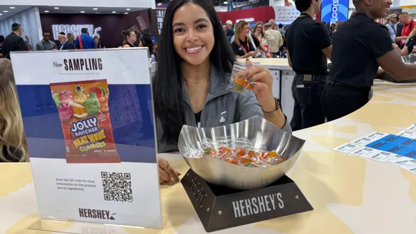 A photo of a eprson holding a small packet of gummies begind a counter. A bowl of gummies sits on the counter. A sign below the bowl says Hershey's and a sign to the side says now sampling Jolly Rancher Heat Wave gummies.