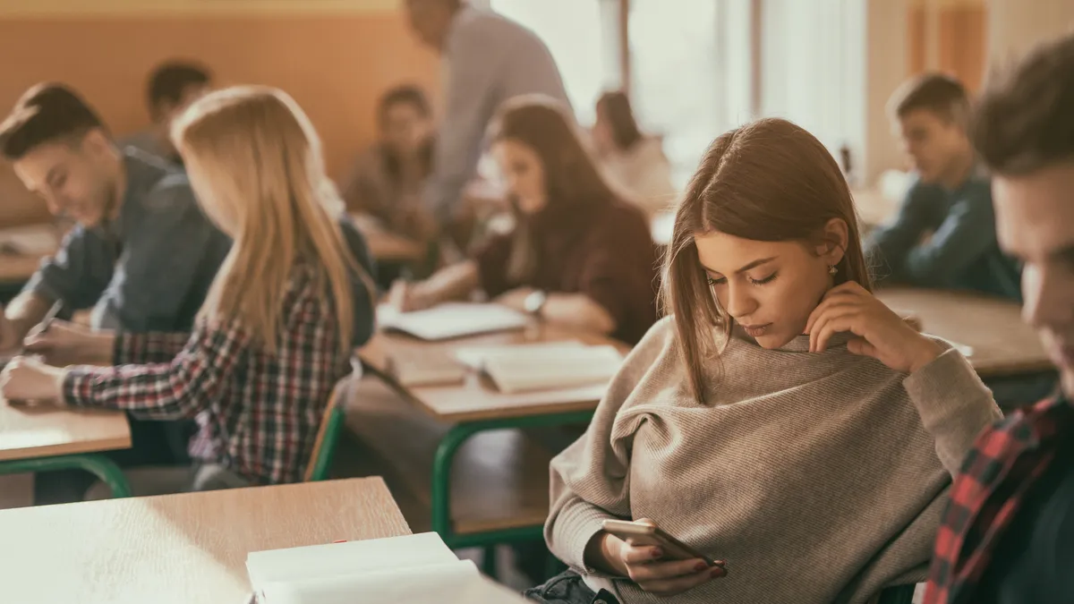 A teenage girl is shown distracted by her smartphone during class time as an educator assists a student in the background.