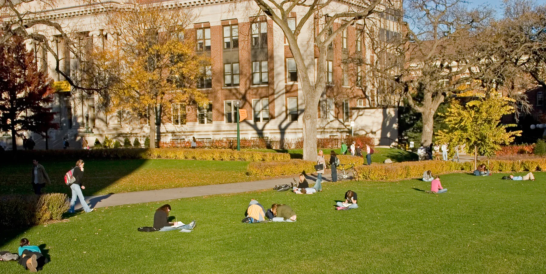 A college quad with students walking and sitting in the grass. Buildings are surrounding the quad.