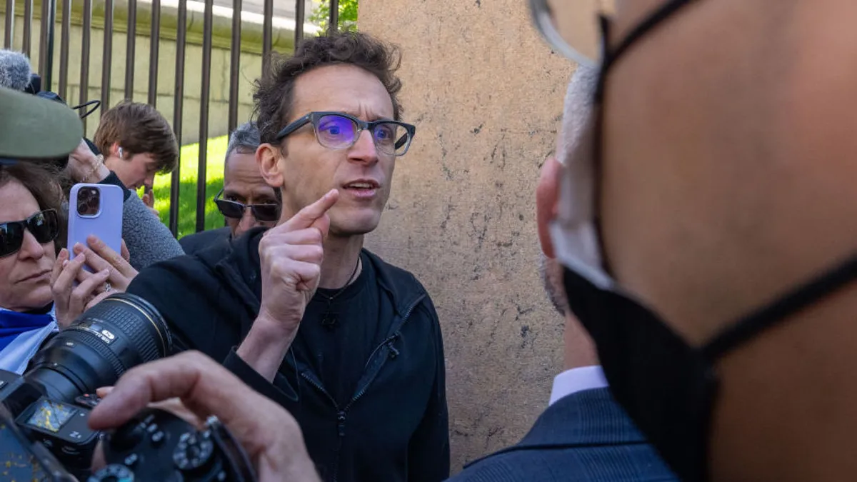 A man in glasses holds up a pointed finger while surrounded by onlookers with cameras.