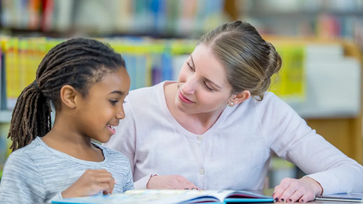 A student and teacher are indoors in a library. They are sitting at a table and reading a book together.