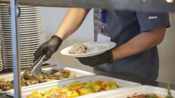 A foodservice employee uses tongs to put food on a plate in a serving line.