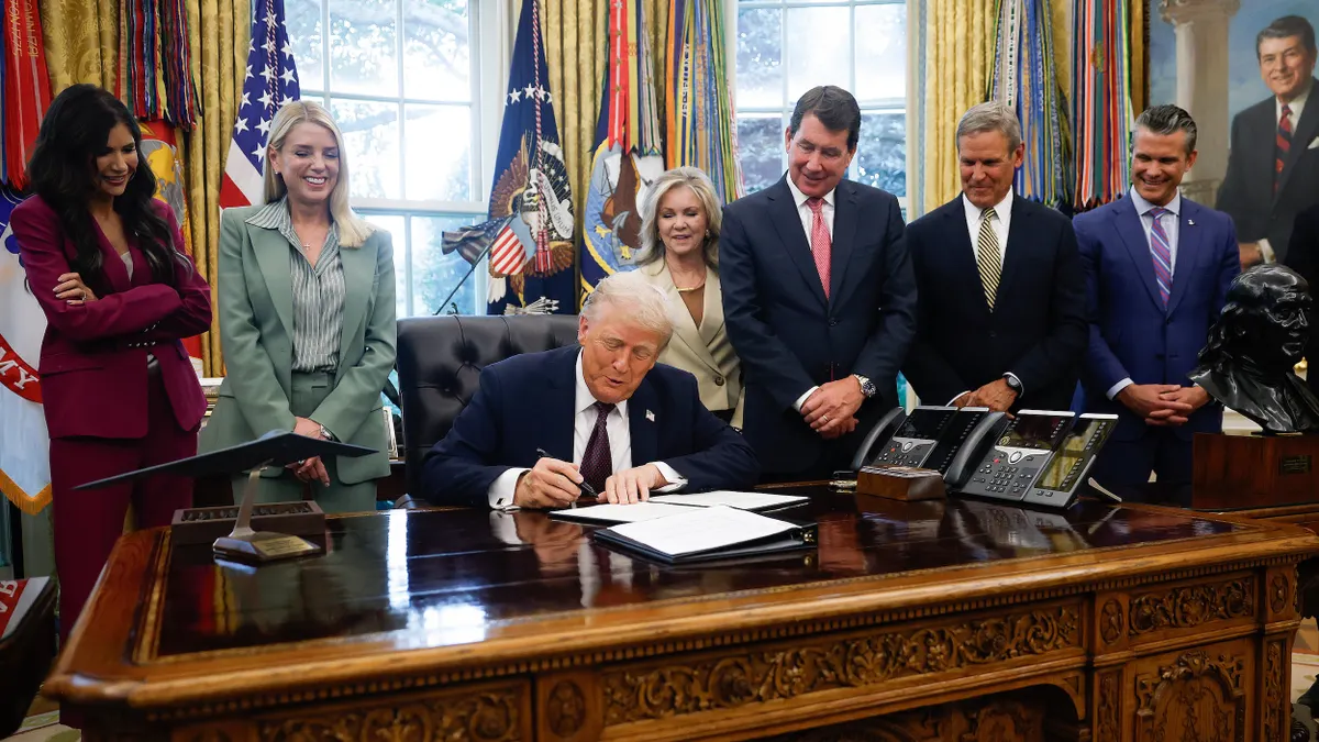 President Trump sits at desk in the Oval Office surrounded by people.