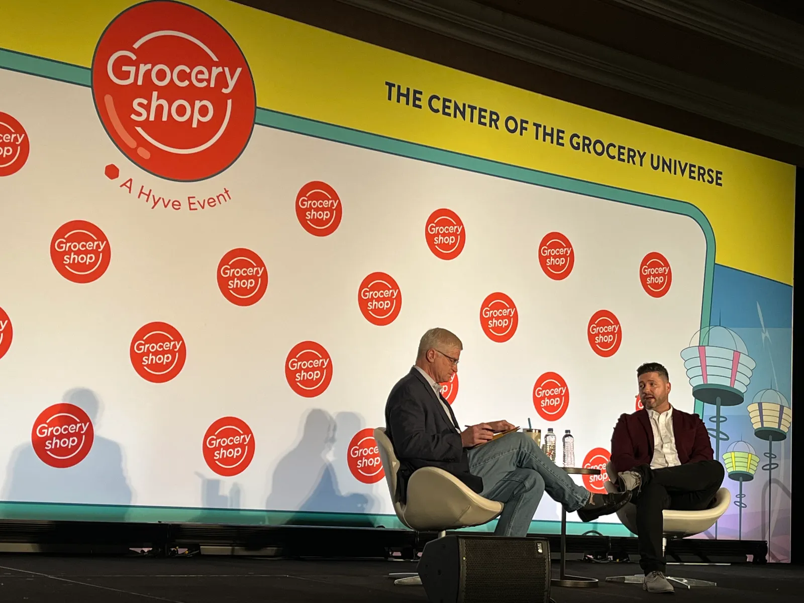 Two people seated on a stage talking to each other against a "Groceryshop" backdrop.