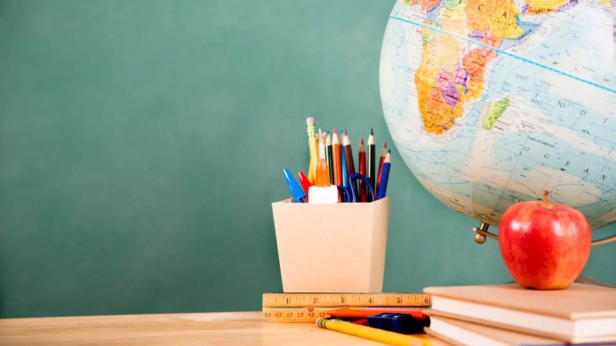 A world globe sits on a desk against a chalkboard background. A red apple, books, ruler and pens and pencils in a pencil holder also sits on the desk.