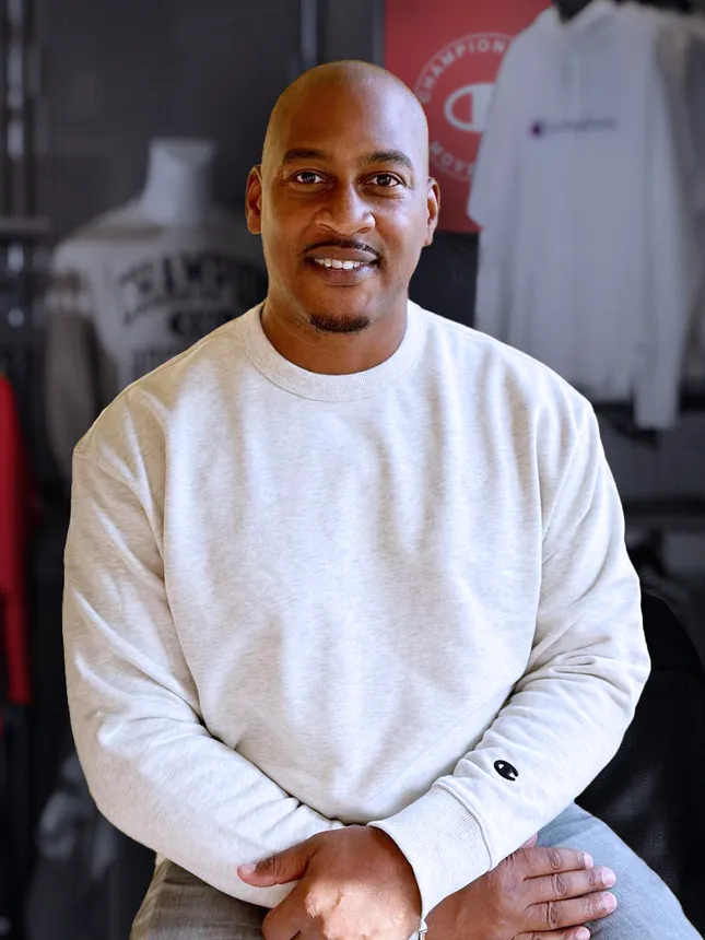 A person sits and smiles at the camera while wearing a gray sweatshirt, and sporting a neatly trimmed facial hair and a cleanly shaved head.