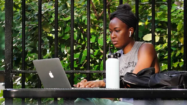 A person sits at a park table, working on a laptop and wearing ear buds.