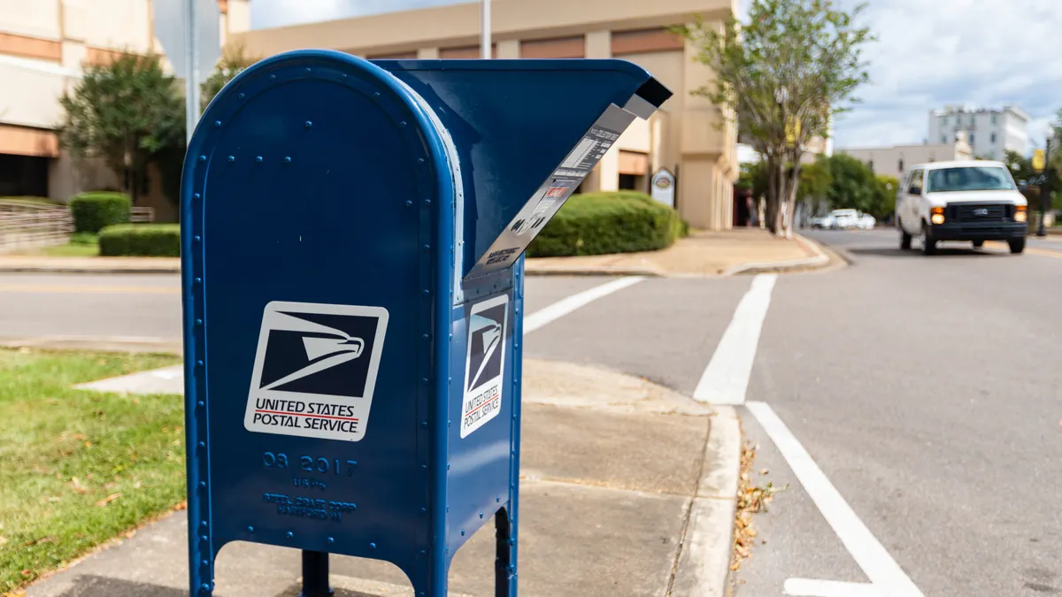 A U.S. Postal Service collection box with a cargo van and street in the background.