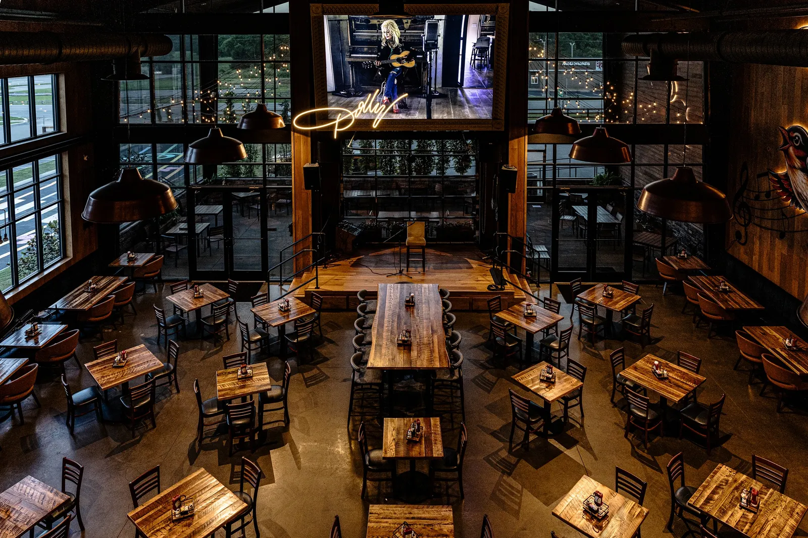 A photo of the dining area inside a large building. The tables are made of metal and wood and a picture of Dolly Parton is on the wall.