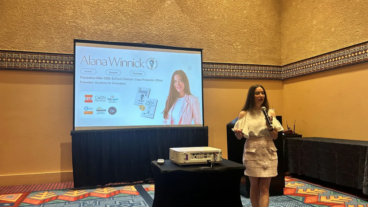A person stands in front of a screen and next to a projector while speaking.
