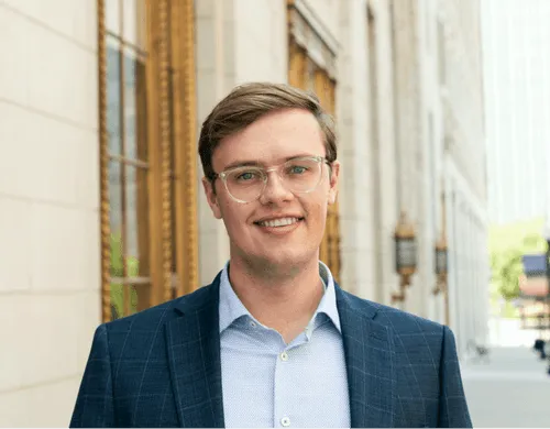 Photograph of a main wearing glasses standing on a sidewalk in front of a building.