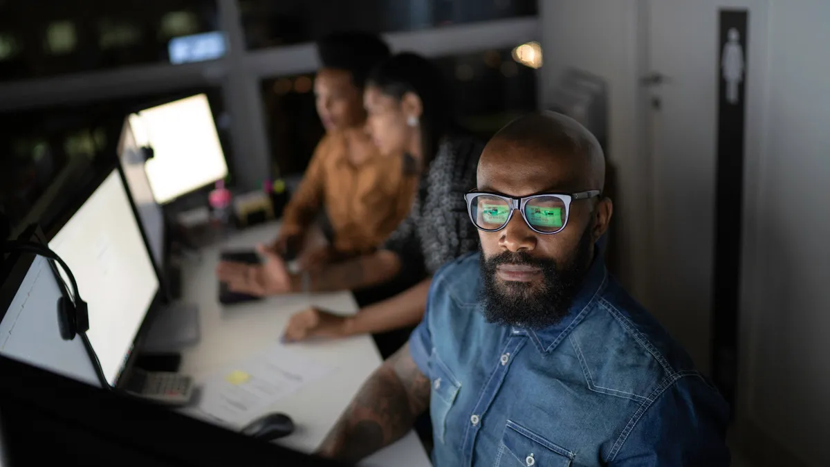 Man working at a computer in a dark office with two other people working behind him out of focus.