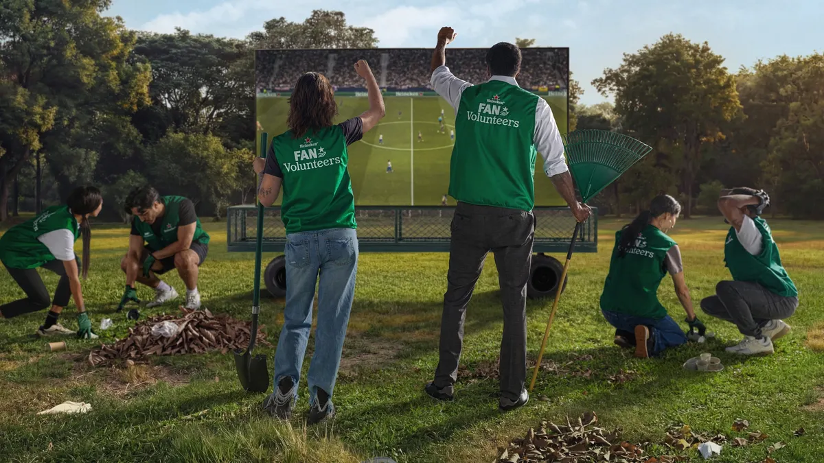 Two people in green vests watch a soccer match as they rake leaves,