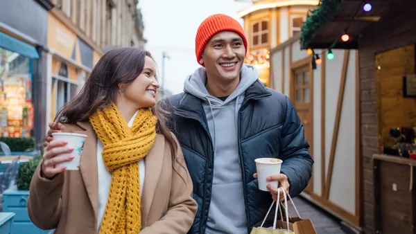 Two people smiling and walking outdoors with coffee cups and shopping bags on a chilly day.