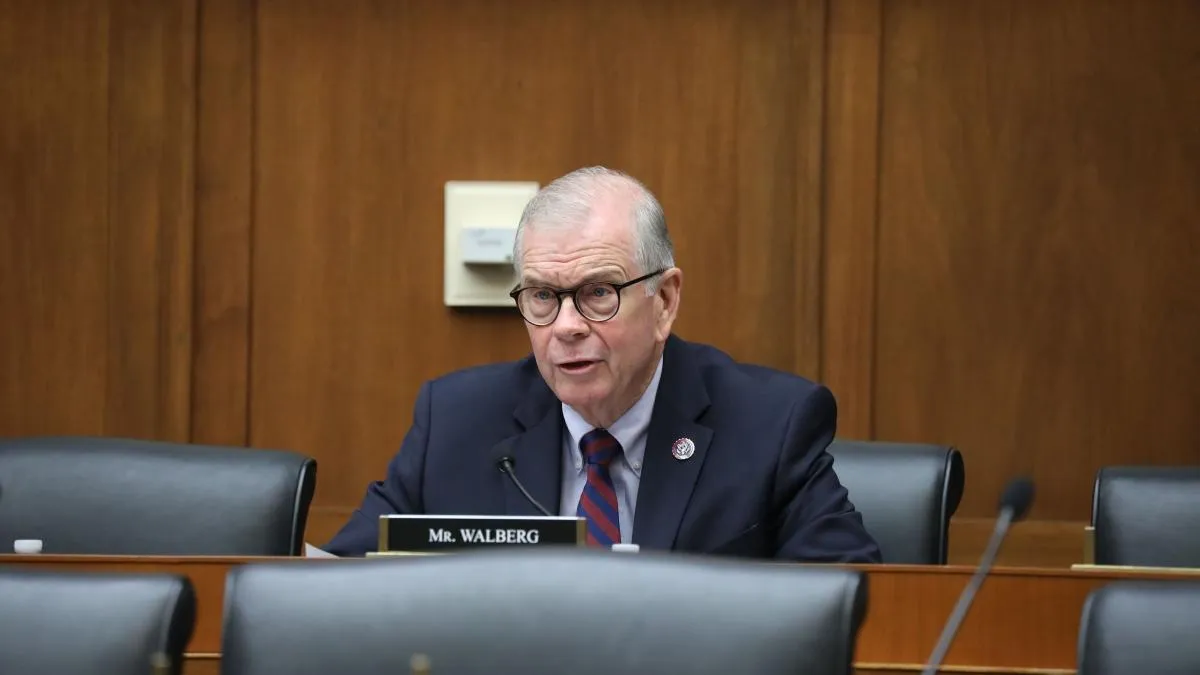 An adult sits in a wood paneled room at a desk. The name plate in front of the person reads "Mr. Walberg"