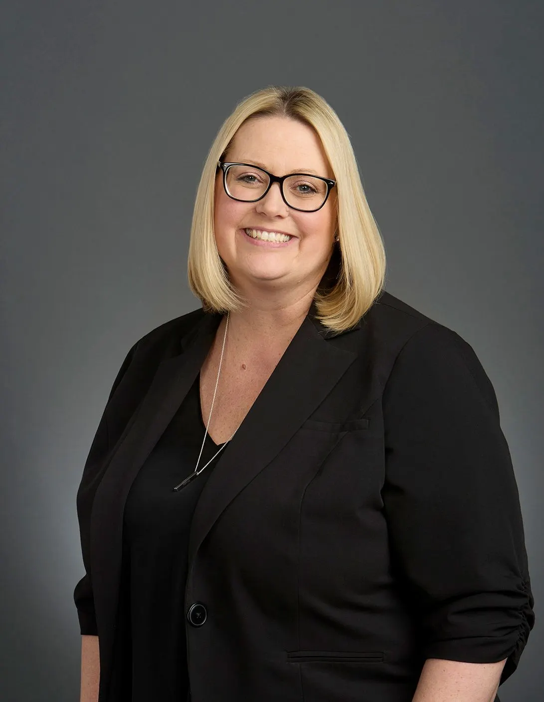 Headshot of a woman with a blonde bob, glasses, against a charcoal gray backdrop