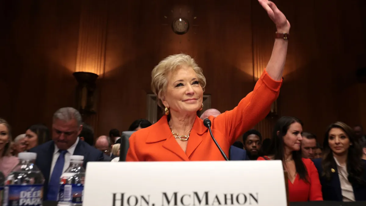 A person is seated as a desk. They are waving their left hand. A name plate sits in front of them and people are seated in row in the background.