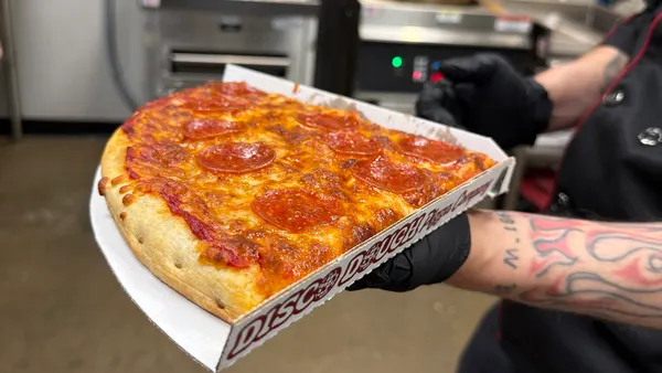 A photo of a person holding a box with a large slice of pizza inside it, standing in a kitchen.