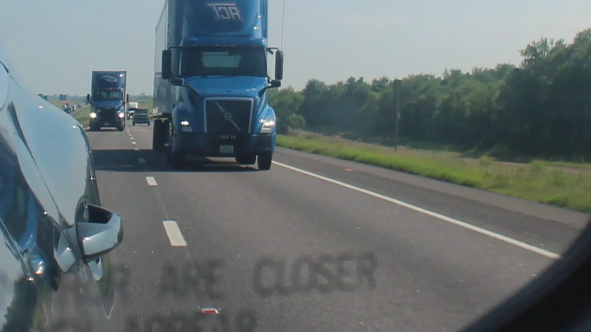 A passenger vehicle mirror shows the front of an ACT tractor driving along a highway.