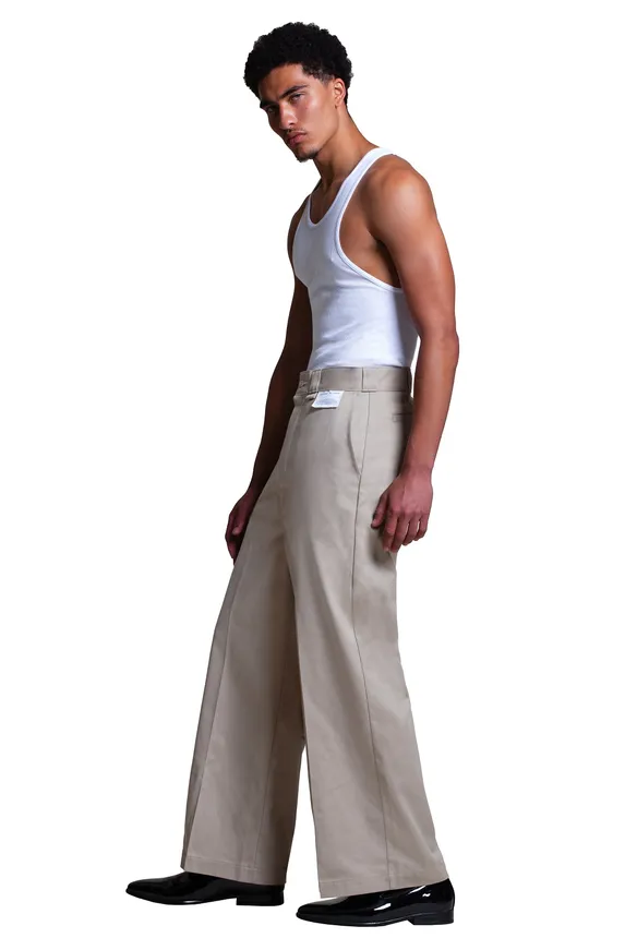 A person stands against a white backdrop wearing Willy Chavarria pants designed for Dickies and a white ribbed tank top.