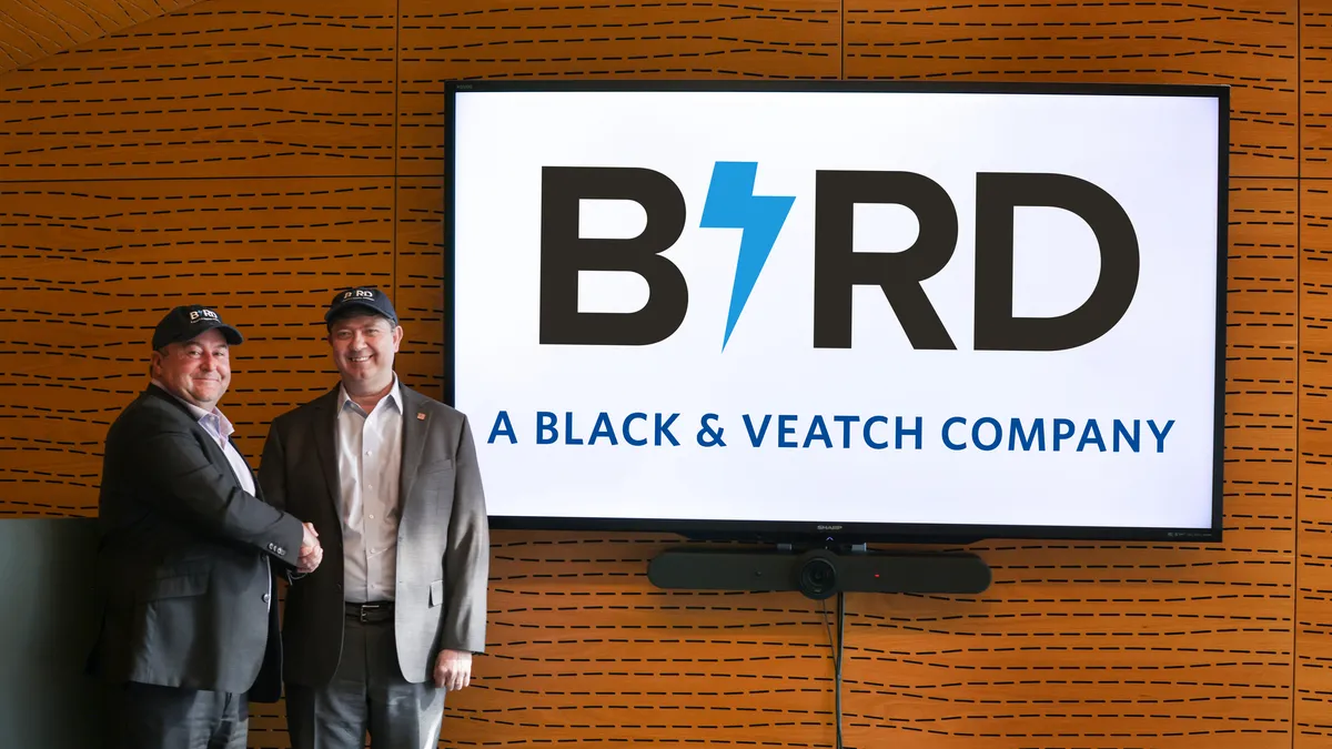 Two men stand next to each other in front of a sign that says "Bird: A Black & Veatch Company" in a wood-paneled room.