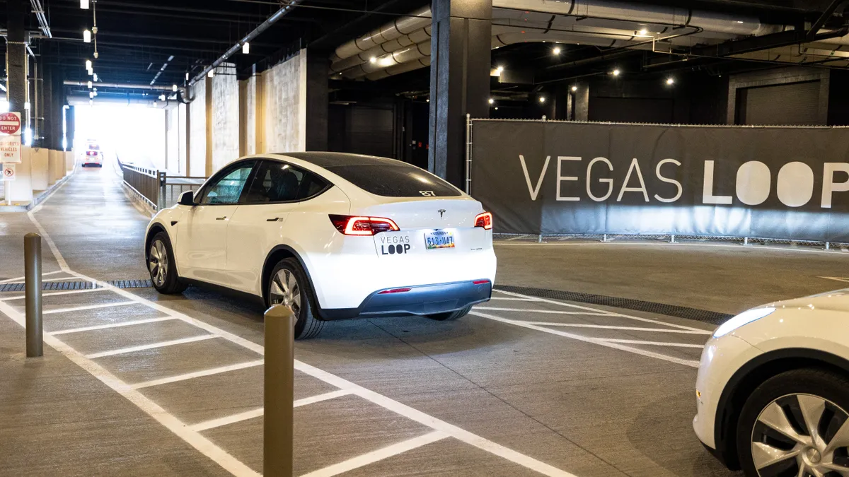 A line of white Tesla Model Ys drives along a narrow road with a Las Vegas Loop in the background.