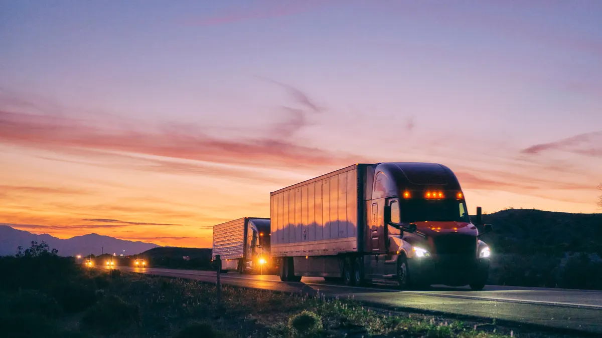 Semis with lights on drive ahead of passenger vehicles in the western U.S. at dusk, with a sunset over mountains.