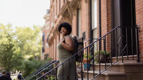 A Black person smiles into the camera; they are wearing a backpack and they are about to commute to work.