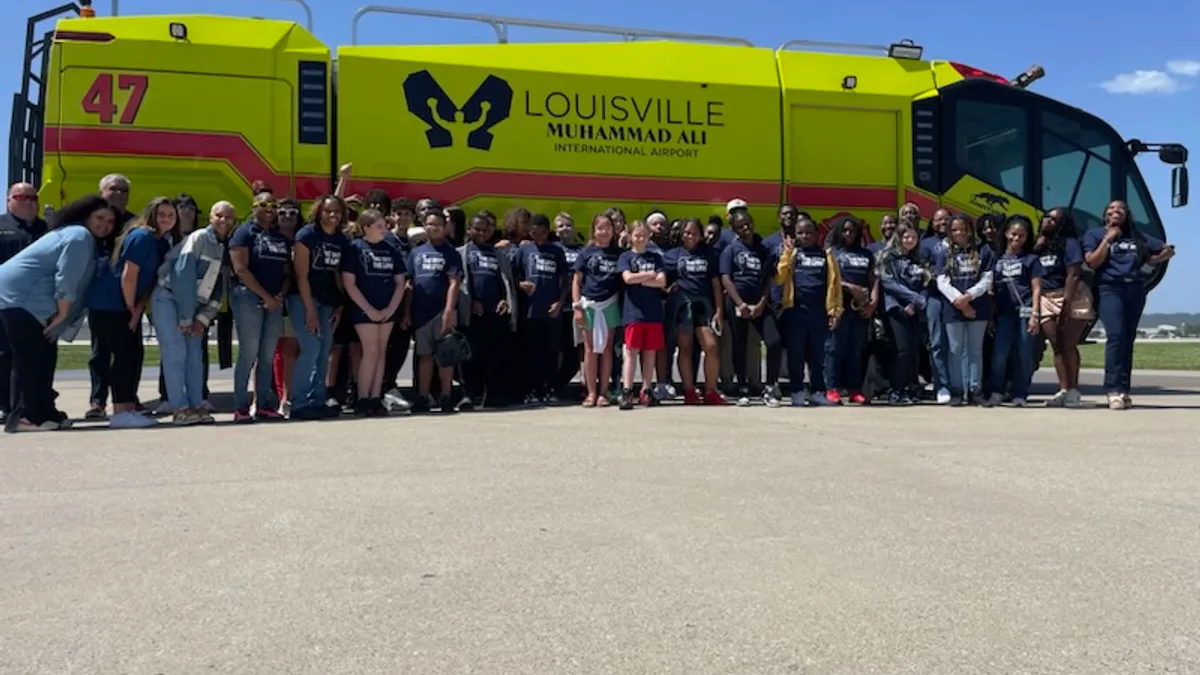 A group of students and adults stand in front of a large vehicle outside at an airport