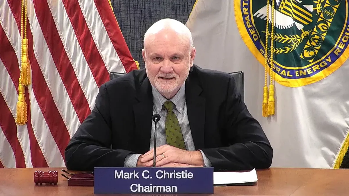 A man sits at a desk in front of a placard with the name Mark Christie, chairman.