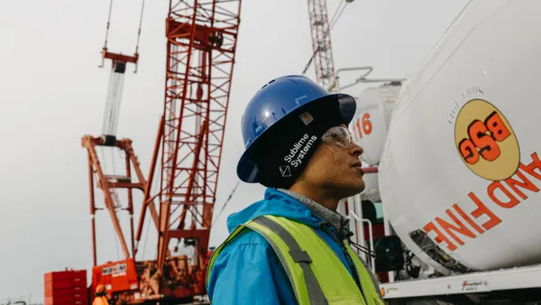 A worker in a blue hard hat and reflective vest stands near a concrete truck, with other construction equipment in the background.