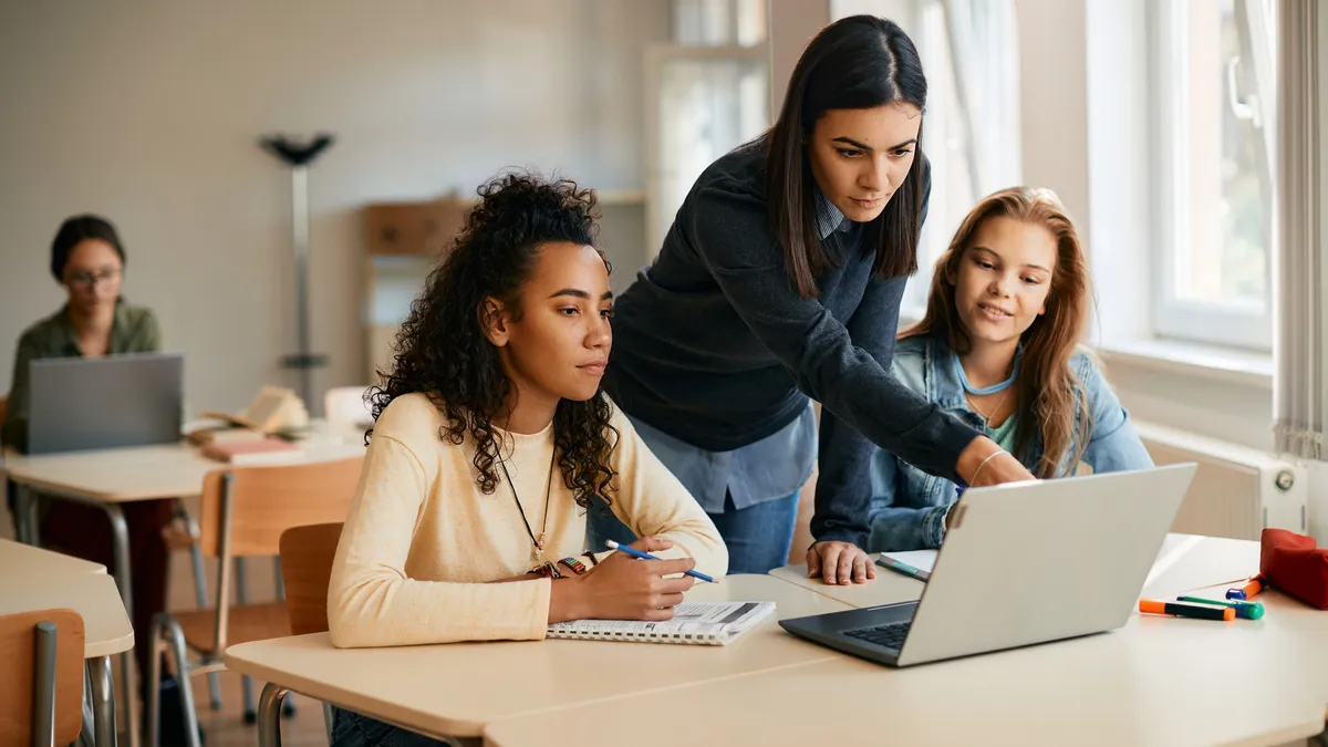 A teacher instructs two students in a classroom while they look onto their laptops.