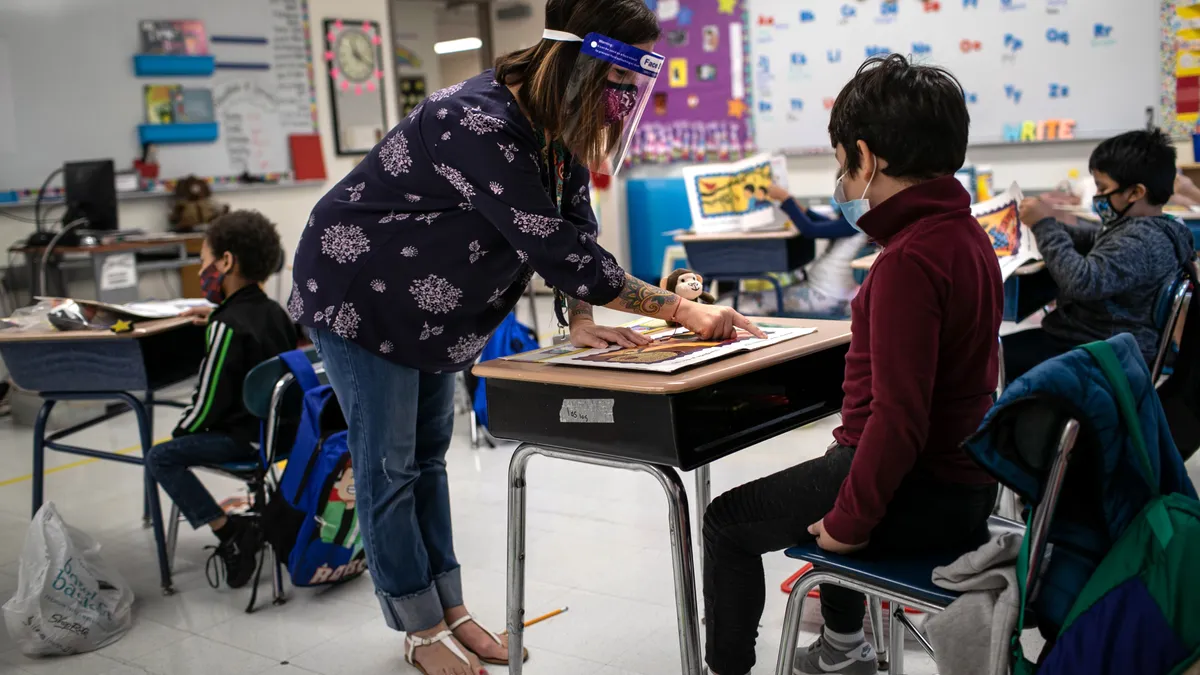 An adult is in a classroom wearing a face mask and shield. Adult is standing in front of a student sitting at a desk.