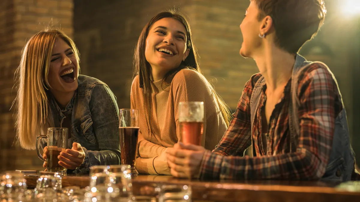 Three cheerful female friends communicating and having fun in a pub during the night out. Focus is on woman in the middle.